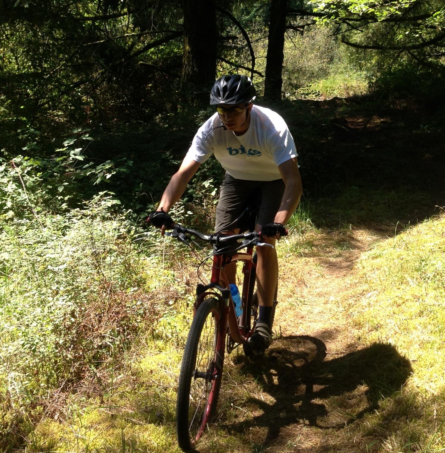 A person riding a mountain bike on a wooded trail, surrounded by greenery. The cyclist is wearing a helmet and a light-colored t-shirt, focused on navigating the path. Sunlight filters through the trees, casting shadows on the ground. Round Lake mountain bike trail.
