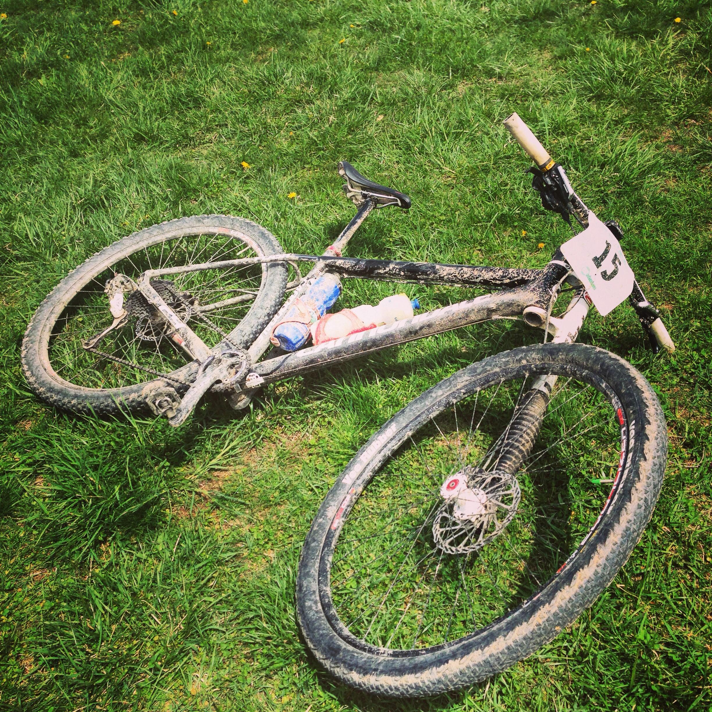 Cannondale Flash 29er 2: A dirt-covered mountain bike lies on grass, featuring a water bottle and a cloth on the frame. The bike has a race number attached to the handlebars, indicating recent use in a race or outdoor event.