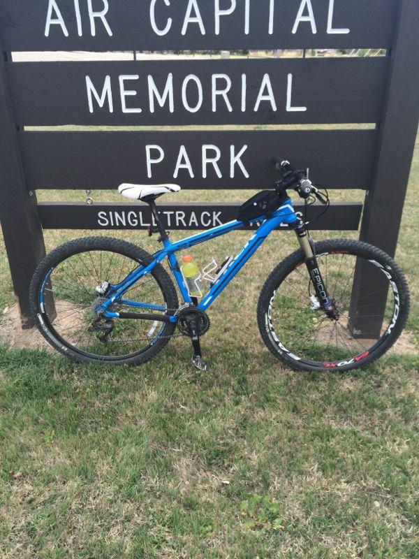 A blue mountain bike parked on grass in front of a sign that reads "Air Capital Memorial Park" with "Single Track" indicated below. The bike has a water bottle attached and is positioned to the left of the sign. Air Capital Memorial Park mountain bike trail.