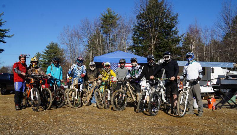 A group of ten people wearing helmets and protective gear stand next to their mountain bikes on a dirt path, with trees and a blue sky in the background. They pose for a photo, showcasing their muddy bikes after a day of riding. A blue tent is set up behind them, contributing to the outdoor atmosphere. Highland Mountain Bike Park mountain bike trail.
