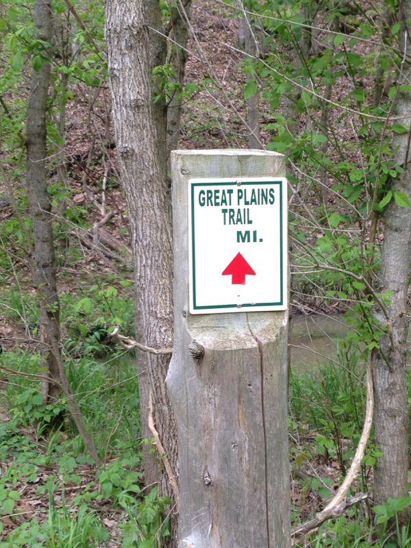 A sign for the Great Plains Trail in Michigan, mounted on a wooden post amidst greenery and trees, featuring an upward arrow indicating the direction of the trail. Indian Camp Creek mountain bike trail.