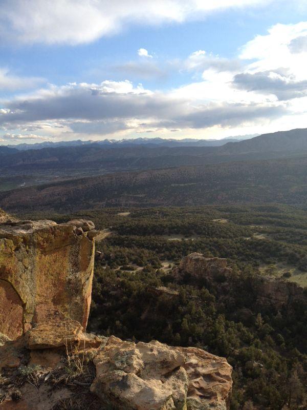 A scenic view from a rocky cliff overlooking a forested valley with distant mountains under a partly cloudy sky. Oil Well Flats mountain bike trail.