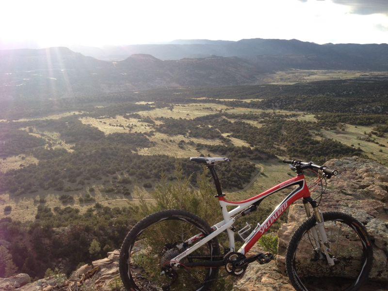Alt tag: A mountain bike parked on a rocky ledge, overlooking a vast landscape of hills and valleys under a cloudy sky with rays of sunlight shining through. Oil Well Flats mountain bike trail.
