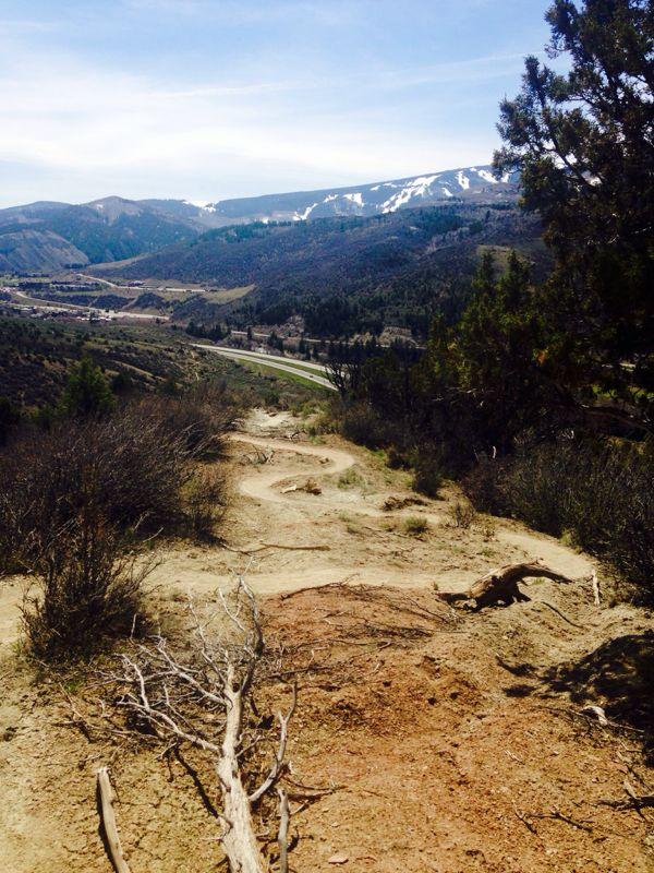 A winding dirt path leads down a hillside, surrounded by sparse vegetation and patches of rocky terrain. In the background, a valley stretches out with a highway visible, bordered by mountains that are partially covered in snow. The sky is clear with a few wispy clouds. Swingletrack mountain bike trail.