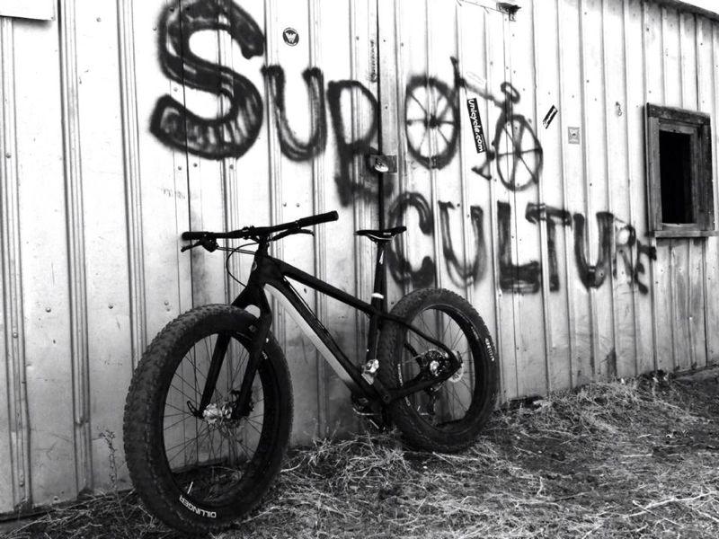 A black and white image of a mountain bike resting against a weathered metal wall. The wall features graffiti that reads "Support Bicycle Culture," along with a bicycle symbol, creating an urban, artistic vibe. The ground is dry, with some grass visible around the bike.