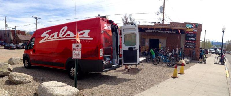 A red Salsa branded van parked outside Subculture Cyclery, with its back doors open. Bicycles are displayed around the entrance of the shop, which features a brick facade and signage. The scene includes a gravel area and a "No Parking" sign, with trees and power lines in the background under a partly cloudy sky.