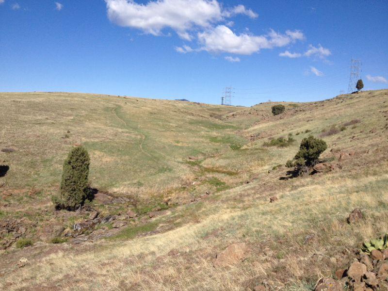 A scenic view of rolling hills covered in dry grass, with a faint walking path winding through the landscape. Sparse trees and rocky outcrops are visible, set against a clear blue sky with scattered clouds. Power lines are seen in the background. North Table Mountain mountain bike trail.