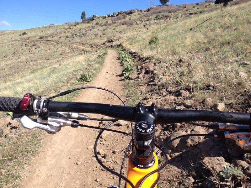 Close-up view of a mountain bike handlebar on a dirt trail, surrounded by grassy hills and rocky terrain. The perspective shows the path ahead winding through the landscape, with a clear blue sky above. North Table Mountain mountain bike trail.
