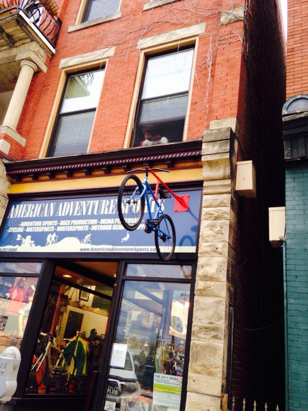 A storefront displays a sign for "American Adventure Sports," featuring a blue and red bicycle mounted on the wall above the entrance. A window above shows a person standing inside, while colorful outdoor gear is visible through the glass. The building is made of red brick, with stone accents and a decorative balcony above.
