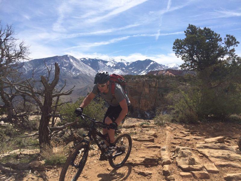A person rides a mountain bike on a rocky trail with a scenic backdrop of snow-capped mountains and a blue sky. The cyclist is wearing a helmet and a backpack, with sparse vegetation and rugged terrain surrounding them. The Whole Enchilada mountain bike trail.