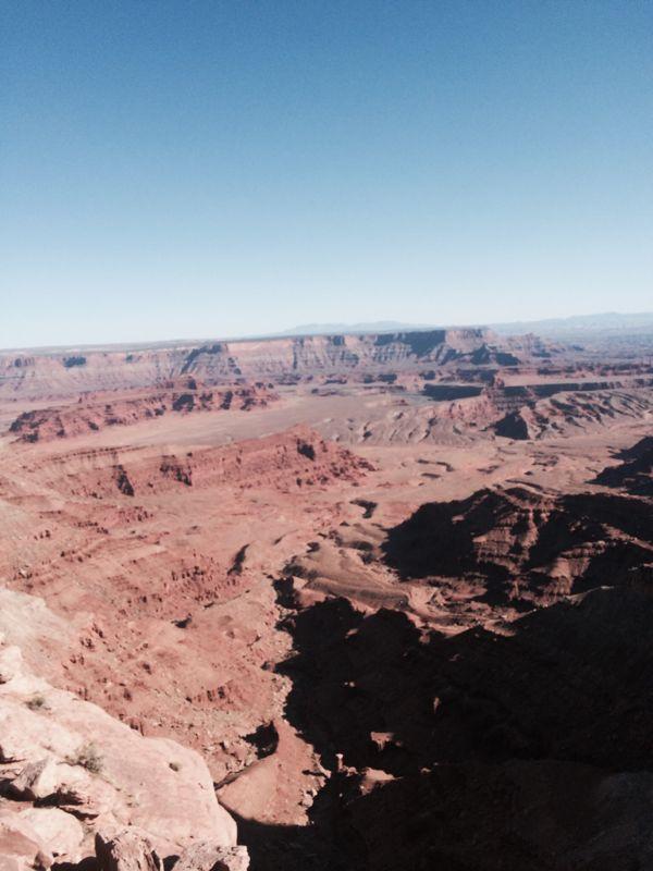 Vast desert landscape featuring layered red rock formations and canyons under a clear blue sky. The scene captures the rugged terrain and expansive views of the canyon's depth and contours. Dead Horse Point State Park mountain bike trail.