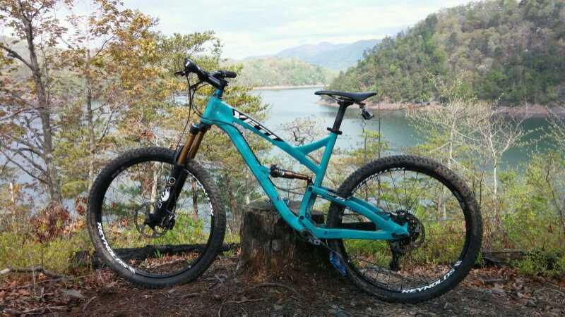 A turquoise mountain bike is propped against a wooden stump, overlooking a scenic lake bordered by trees and rolling hills in the background. The peaceful landscape features a calm, reflective water surface under a partly cloudy sky. Tsali Thompson Loop mountain bike trail.