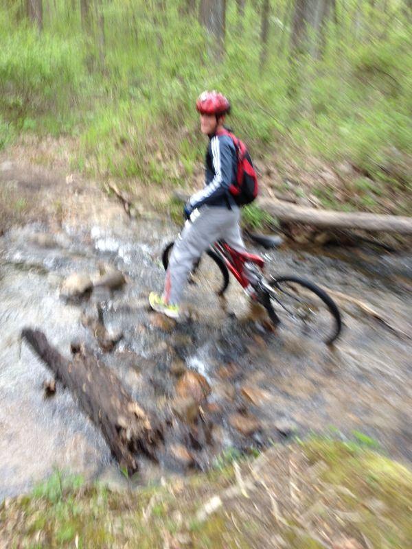 A person riding a mountain bike across a rocky stream in a wooded area. The cyclist is wearing a red helmet and a backpack, and is navigating over stones and logs in the water. The surrounding landscape features green foliage and trees. The image appears slightly blurred, suggesting movement. Greenbrier State Park mountain bike trail.