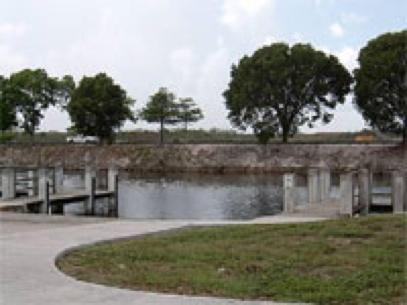 A peaceful landscape featuring a curved concrete path leading towards a body of water, bordered by several trees. Wooden docks are visible along the water's edge, set against a cloudy sky. Markham Park mountain bike trail.