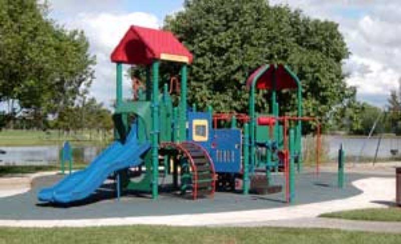A colorful playground featuring a blue slide, climbing structures, and a tunnel, surrounded by green grass and trees, with a pond in the background under a partly cloudy sky. Markham Park mountain bike trail.