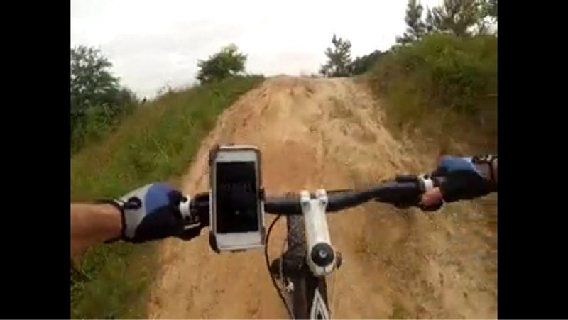 A cyclist's perspective on a bike trail, showing the handlebars with a phone mounted on the left side, navigating a sandy incline surrounded by greenery. Huntsville State Park mountain bike trail.