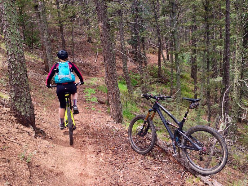 A person riding a mountain bike on a dirt trail through a forest, with tall trees and greenery surrounding the path. A second mountain bike is parked nearby. The rider wears a helmet and a colorful backpack. Cheyenne Mountain State Park mountain bike trail.