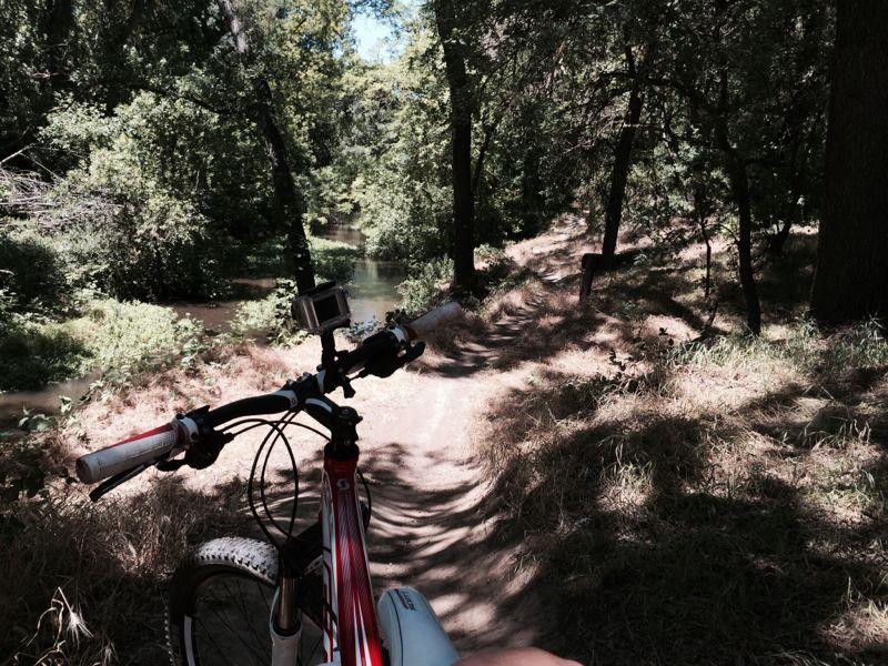 A mountain bike parked on a dirt path surrounded by lush greenery, with a river visible in the background. Sunlight filters through the trees, creating dappled shadows on the ground. The bike's handlebars and part of a person's leg are in the foreground, emphasizing a peaceful outdoor setting. Dry Creek Trail mountain bike trail.