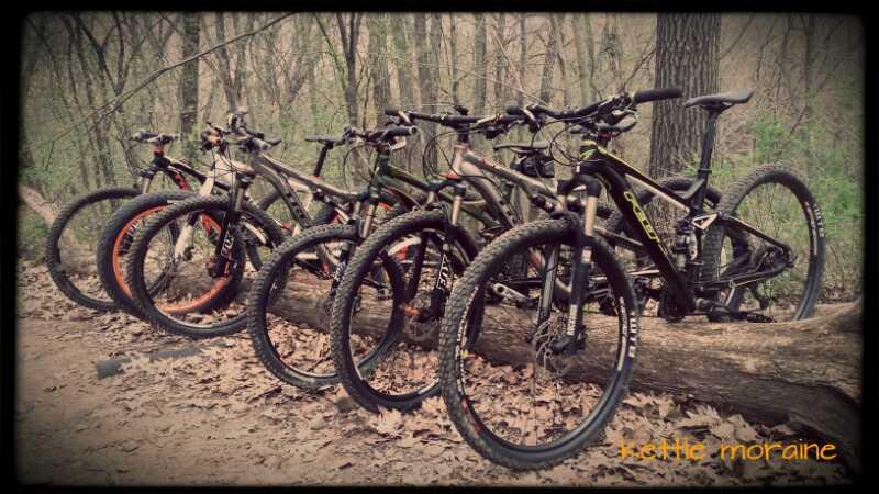A row of mountain bicycles parked against a fallen log in a wooded area, surrounded by a carpet of leaves. The bicycles have various frame colors and are equipped with knobby tires, indicating they are suited for off-road riding. The scene captures a tranquil moment in nature, highlighting the connection between cycling and the outdoors. Kettle Moraine John Muir + Emma Carlin mountain bike trail.
