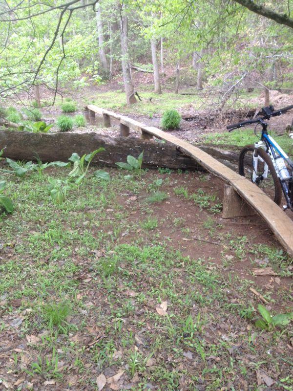 A wooden bike bridge extends over a small pathway in a wooded area, with greenery and small plants surrounding it. A mountain bike is leaning against the side of the bridge. The scene is lush and tranquil, suggesting a nature trail. Allaire State Park mountain bike trail.
