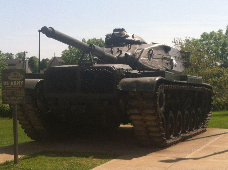 A Soviet-era tank displayed outdoors on a concrete surface, with a sign nearby indicating it is part of a military exhibit. The tank is positioned at an angle, showcasing its large turret and tracks, and is surrounded by greenery and houses in the background under a clear blue sky. Veterans Park mountain bike trail.
