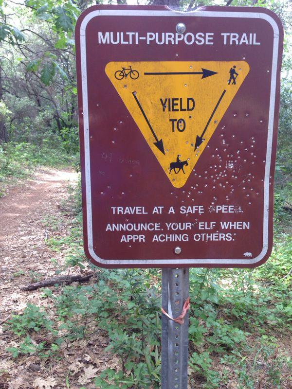 A weathered sign at the edge of a multi-purpose trail, featuring directional arrows for cyclists, pedestrians, and horseback riders. The sign instructs users to yield to others and includes humorous typos, such as "travel at a safe pee" and "announce your 'elf' when approaching others." Greenery surrounds the sign, indicating a natural outdoor setting. Clementine / Forresthill Connector Trail mountain bike trail.