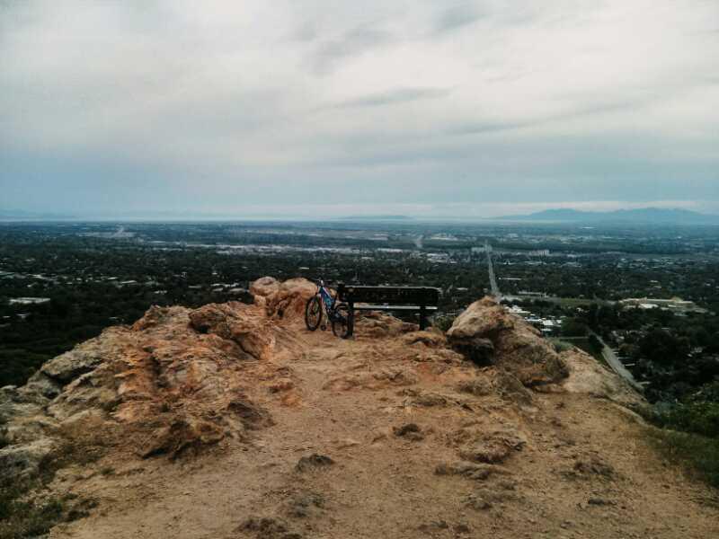 A mountain bike rests on a rocky outcrop overlooking a vast landscape, with a wooden bench beside it. The view includes a mix of urban and green areas below, under a partly cloudy sky. Bonneville Shoreline mountain bike trail.