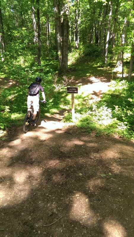 A person on a mountain bike rides along a dirt trail in a lush green forest. In the foreground, a caution sign is visible, warning about a steep incline ahead. Sunlight filters through the trees, casting dappled shadows on the ground. Schaeffer Farms mountain bike trail.