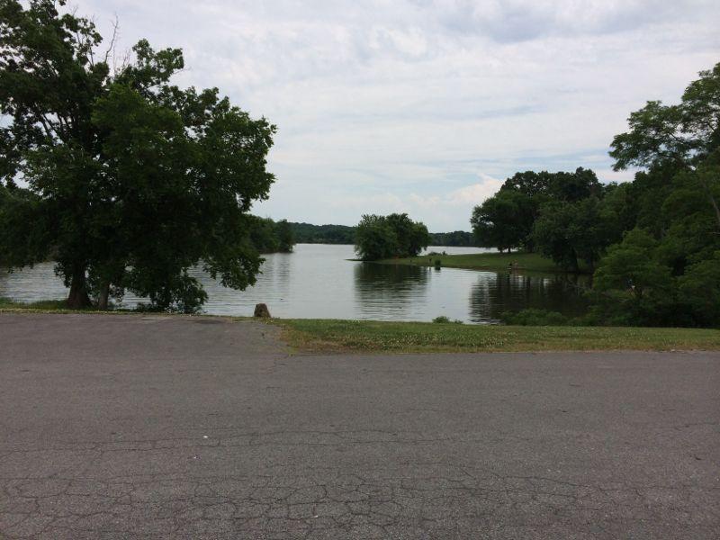 A serene view of a calm lake surrounded by lush greenery, with trees lining the shore. The sky is partly cloudy, reflecting soft light onto the water's surface, creating a peaceful and relaxing atmosphere. Lock 4 mountain bike trail.