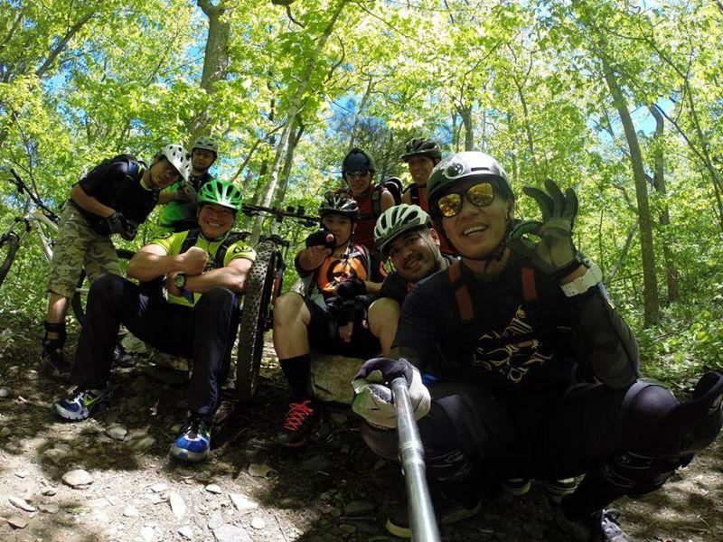 A group of nine people posing together for a selfie in a lush green forest. They are dressed in biking gear and wearing helmets, with bicycles nearby. The atmosphere is cheerful, capturing a moment of camaraderie during an outdoor biking adventure. Sunlight filters through the trees, adding a warm glow to the scene. Rocky Knob Park mountain bike trail.