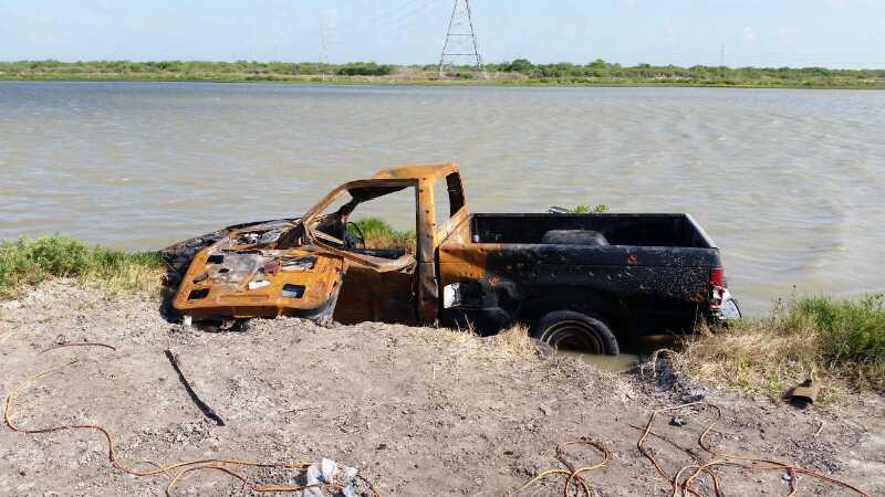 An abandoned and rusty pickup truck partially submerged near a body of water, surrounded by grass and dirt. Power lines are visible in the background. The truck shows signs of decay and neglect. Oso Creek Park mountain bike trail.