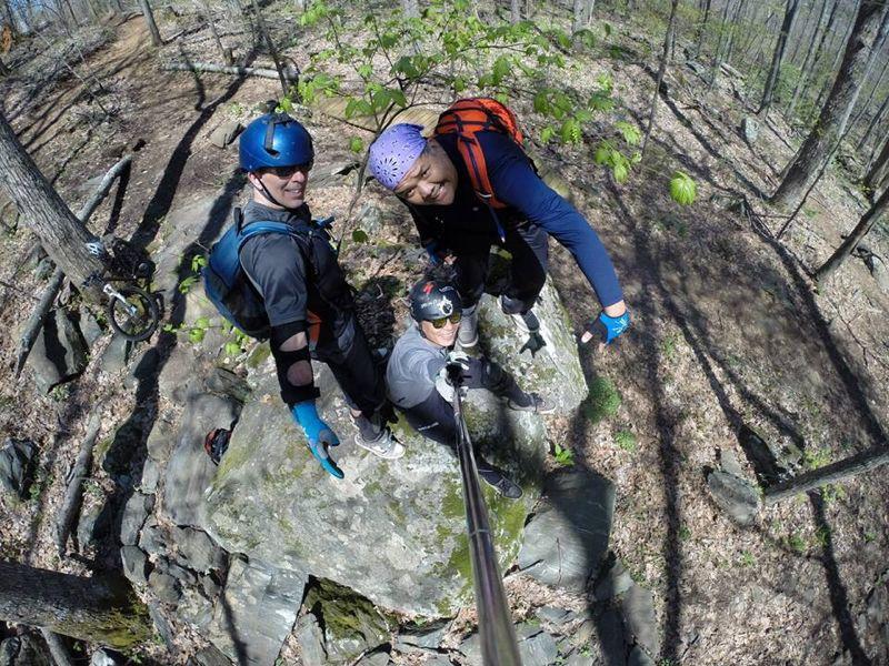 Three people take a selfie while standing on a rock in a forested area. They are wearing helmets and outdoor gear, surrounded by trees and rocks. The scene is bright and captures the essence of an outdoor adventure. Rocky Knob Park mountain bike trail.