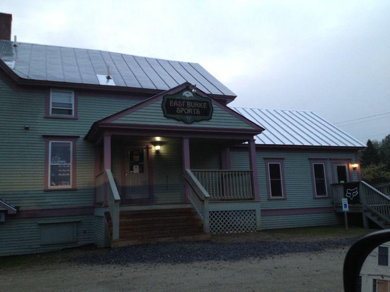 A wooden building with a green exterior and purple trim, featuring a metal roof. The sign above the entrance reads "East Burke Sports." The front has a porch with steps leading up to it, and there are lights visible on the building. The scene is captured during twilight, creating a soft, muted atmosphere. A parked vehicle is partially visible in the foreground.