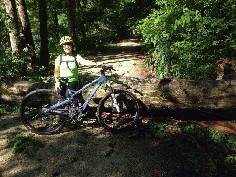 A person wearing a bright green shirt and a helmet stands beside a blue mountain bike on a forest trail, with a fallen log blocking part of the path. The scene is surrounded by lush greenery, showcasing a sunny day in nature. Salem Lake mountain bike trail.