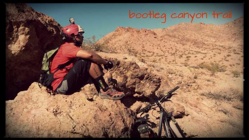 A person wearing a red shirt, black shorts, and a helmet sits on a rocky outcrop along Bootleg Canyon Trail, with a mountain bike resting beside them. The background features a desert landscape with rolling hills and sparse vegetation under a clear blue sky. Bootleg Canyon mountain bike trail.