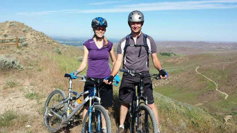 A woman and man standing beside their mountain bikes on a hillside with a scenic view. The woman is wearing a purple shirt and blue helmet, while the man is in a gray shirt and helmet. The landscape features rolling hills and a clear blue sky. Kestrel, Sidewinder, Hulls Gultch mountain bike trail.