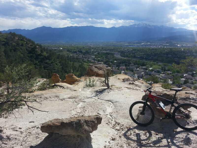 A panoramic view from a rocky outcrop overlooking a valley and mountains in the distance, with a mountain bike parked nearby. The landscape features greenery, houses, and winding roads below, under a partly cloudy sky. Palmer Park mountain bike trail.