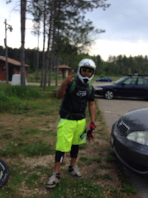 A person wearing a motocross helmet, gloves, and a black shirt is giving a peace sign while standing in a grassy area. They are dressed in bright neon yellow shorts and have knee pads on. In the background, there are trees and parked cars. Emma Carlin mountain bike trail.