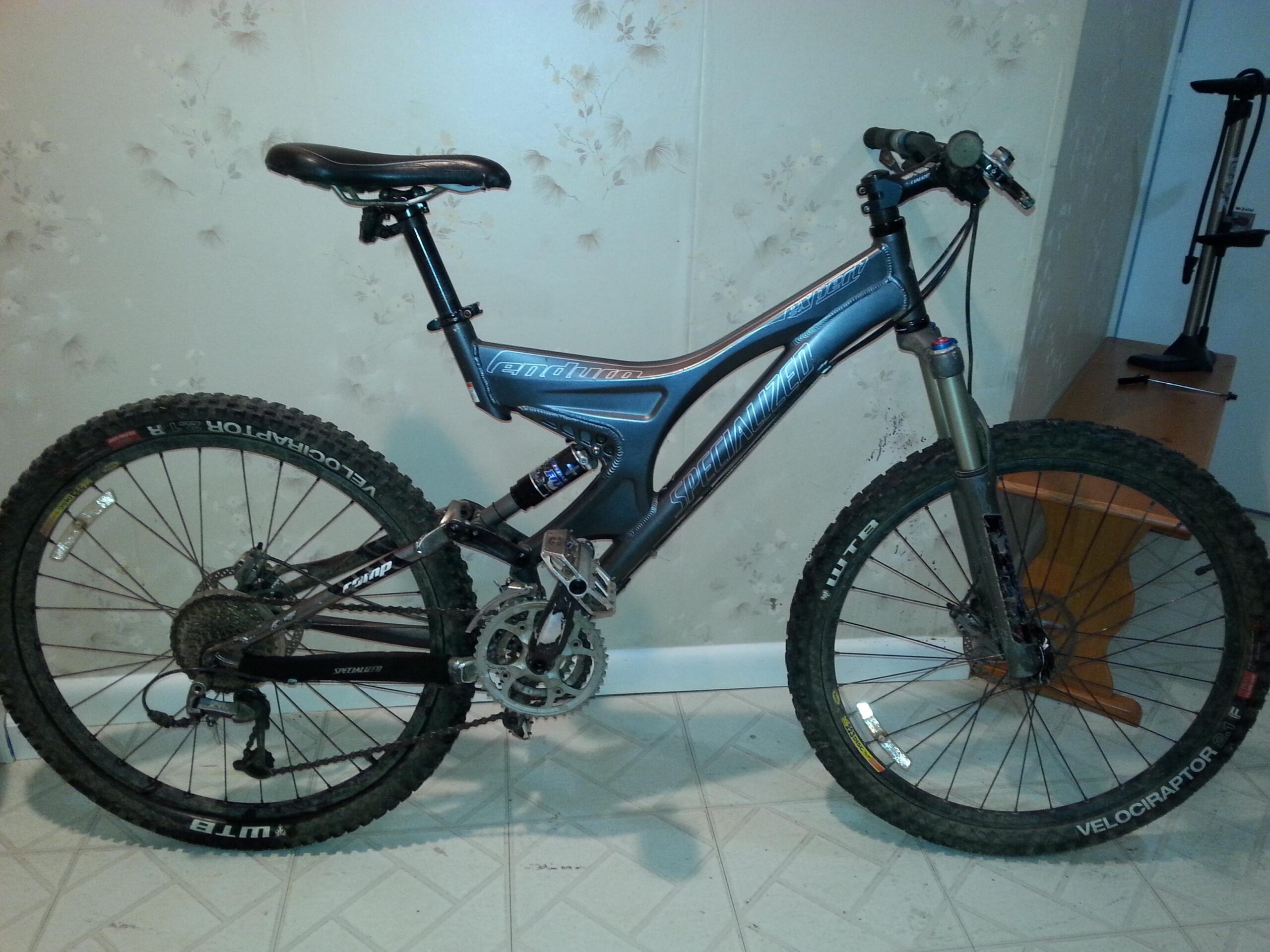 Specialized Enduro Expert: A mountain bike is positioned against a light floral-patterned wall. The bike features a sleek gray frame with the name "Specialized" prominently displayed. It has thick tires designed for off-road use, with the brand "WTB Velociraptor" visible on the tire sidewalls. The bike's seat is raised, and it has a suspension fork at the front. A floor pump is partially visible in the background, along with a wooden table.