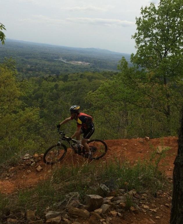 A mountain biker maneuvering along a dirt trail on a hillside, surrounded by lush green trees and a distant view of rolling hills under a partly cloudy sky. Coldwater Mountain mountain bike trail.