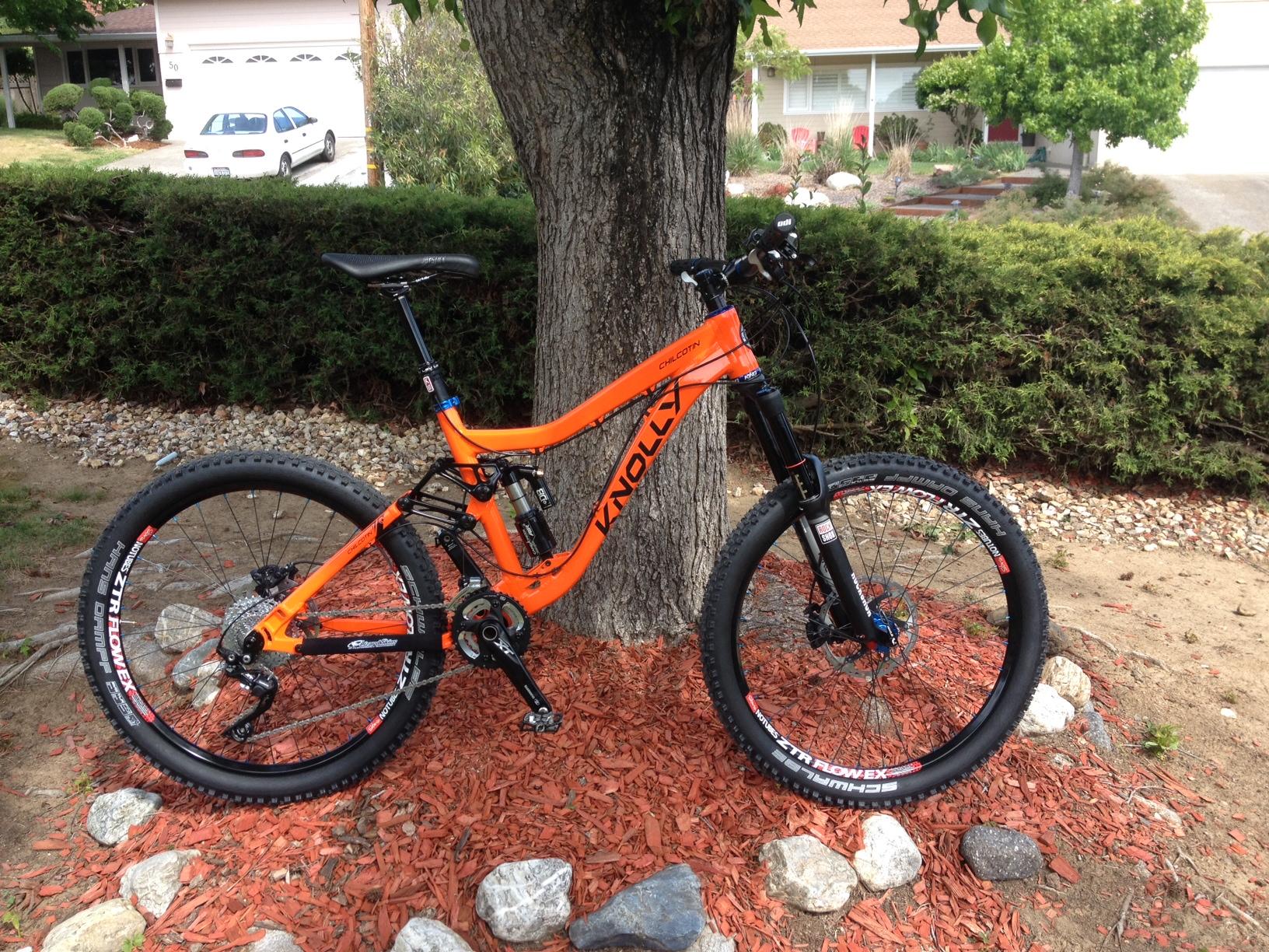 Knolly Chilcotin: An orange Knolly mountain bike stands against a tree in a landscaped yard, surrounded by mulch and stones. In the background, a house with a driveway is partially visible. The bike features wide tires, a sturdy frame, and suspension components, indicating it is designed for off-road riding.
