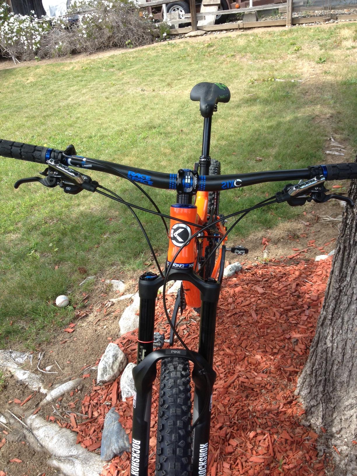 Knolly Chilcotin: A close-up view of a mountain bike's handlebars, showcasing the orange frame, black handlebars, and various components including brake levers and a gear shifter. The bike is positioned on a grassy area with reddish-brown mulch in the foreground and some rocks visible. In the background, there are trees and a wooden structure partially obscured by foliage.