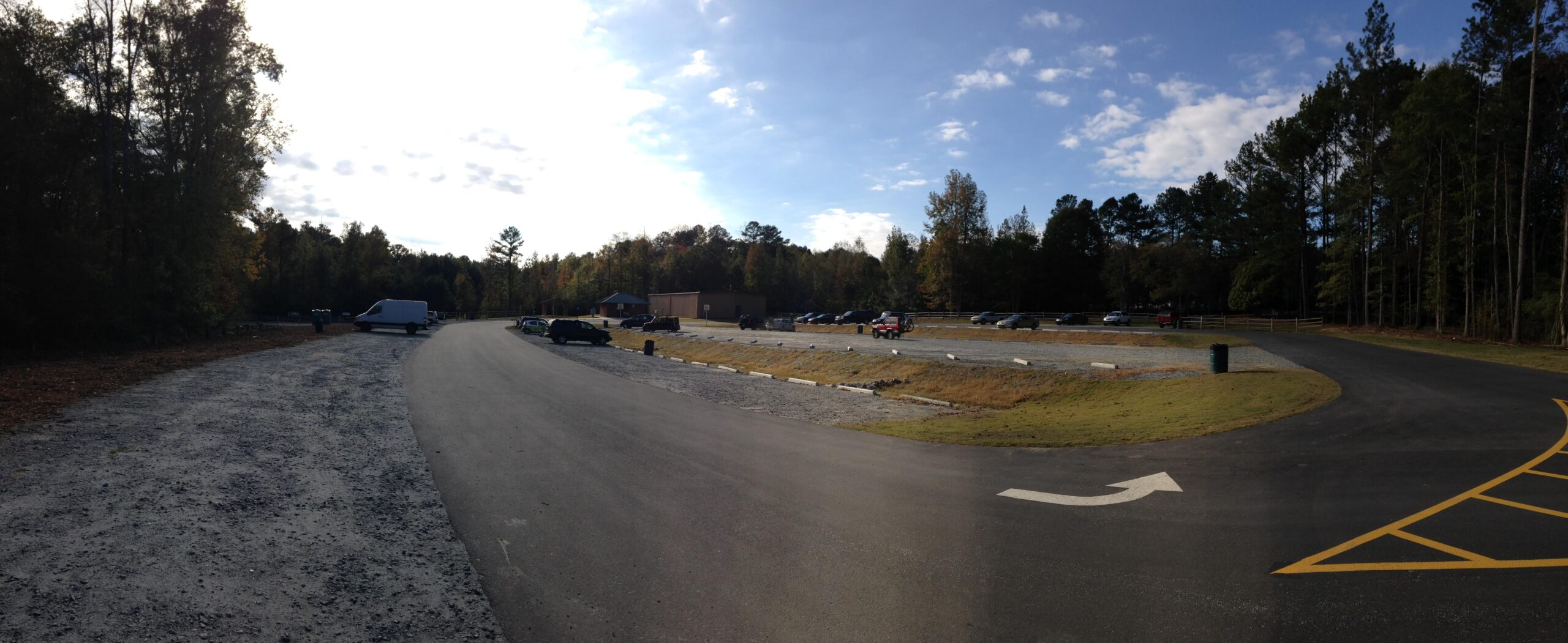 A panoramic view of a parking lot surrounded by trees, showcasing a variety of parked vehicles, including cars and a van. The parking lot features both paved and gravel areas, with a road curving towards the right. In the background, there is a building partially obscured by trees, under a partly cloudy sky. Blankets Creek mountain bike trail.