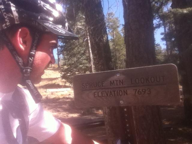 A person wearing a cycling helmet is closely examining a wooden sign that reads "Spruce Mtn. Lookout" and notes the elevation as "7693." The surrounding area features tall trees, indicating a forested environment. Lynx Lake And Spruce Mountain Loop mountain bike trail.