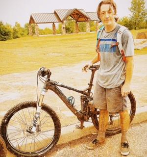 Trek Fuel EX 8: A young person standing next to a mountain bike on a gravel path, wearing a gray t-shirt and shorts, with mud on their clothing and bike tires. In the background, there are trees and a pavilion. The sky is clear and sunny.