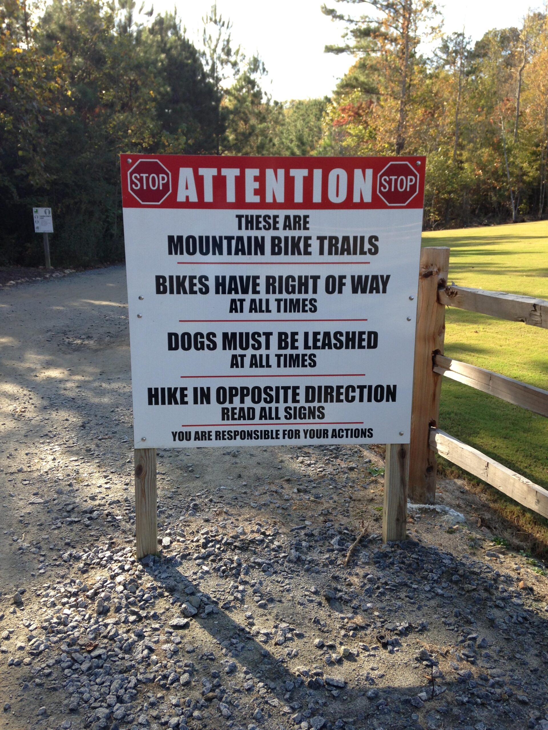 A sign at the entrance of a mountain bike trail, featuring important regulations: "Attention: These are mountain bike trails. Bikes have right of way at all times. Dogs must be leashed at all times. Hike in opposite direction. Read all signs. You are responsible for your actions." The setting includes a dirt path and surrounding trees. Blankets Creek mountain bike trail.