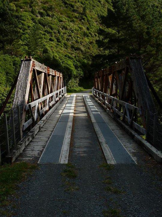 A rustic wooden bridge spans a narrow path, surrounded by lush greenery. The bridge features a slatted design with a mix of gray and weathered wood, casting long shadows across the walkway. Sunlight filters through the trees, highlighting the bridge