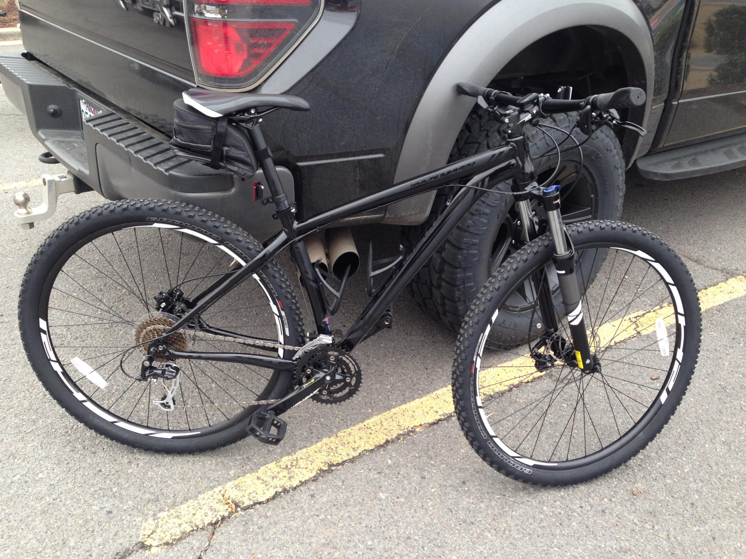 Specialized Rockhopper 29: A black mountain bike parked next to a large pickup truck. The bike's tires are suitable for off-road terrain, and it features a modern design with visible gear components. The background shows a portion of the truck and the asphalt surface of a parking lot.