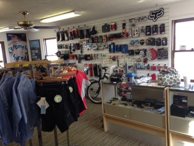 A view of the interior of a retail store featuring bike accessories and apparel. Displayed are various items on the walls, including helmets, tools, and gear, alongside hung shirts and shorts in the foreground. A bicycle is visible in the background, and a glass display case showcases additional products. Natural light comes through the windows, illuminating the space.