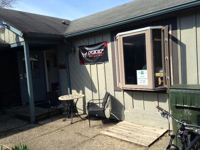 Exterior view of a small bike shop featuring a sign that reads "Great Bicycles." The entrance has a table and chairs set up outside, along with a recycling bin nearby. A bicycle is parked against the building. The shop has light green wooden siding and a large window that is partially open. The scene is set on a sunny day.